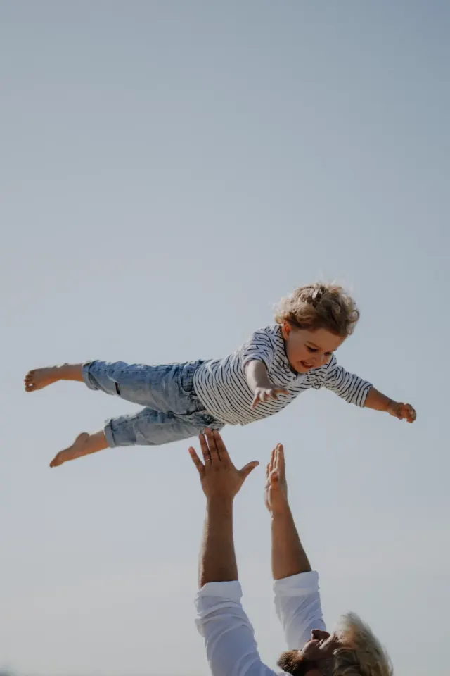 Petit garçon lancé dans les airs par son père, bras grands ouverts, séance famille sur la plage de Leffrinckoucke