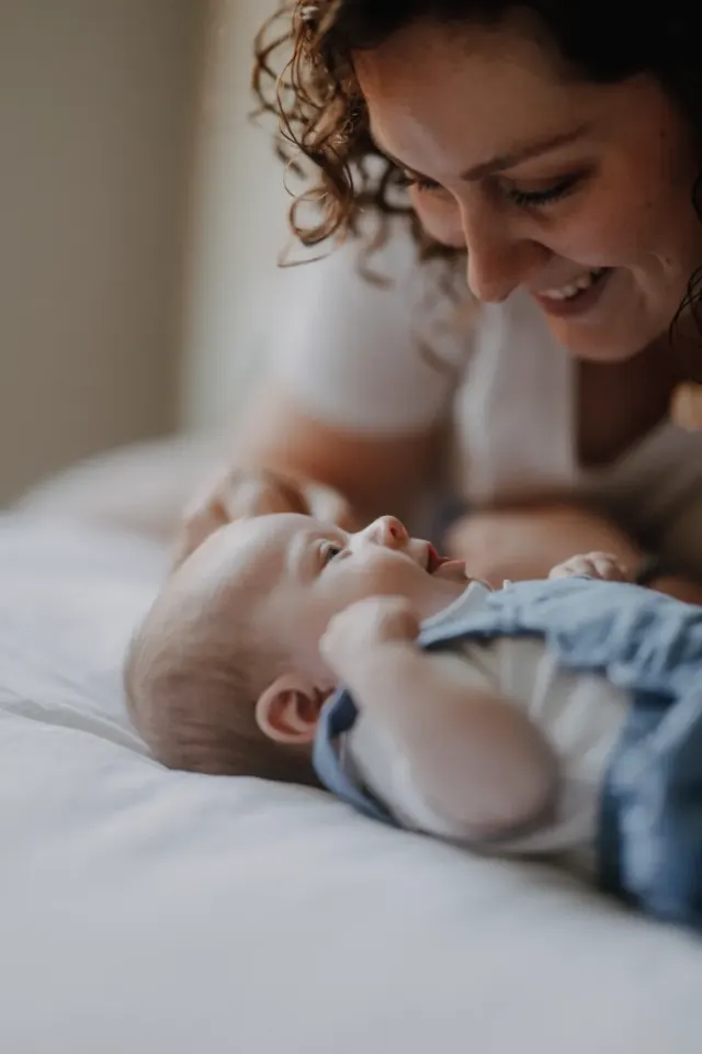 Maman penchée en souriant sur son bébé allongé sur le lit, séance naissance à domicile à Auchy-lez-Orchies