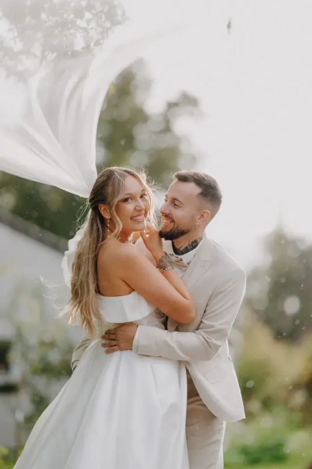 Mariés enlacés et rieurs, le voile soulevé par le vent, séance couple le jour du mariage