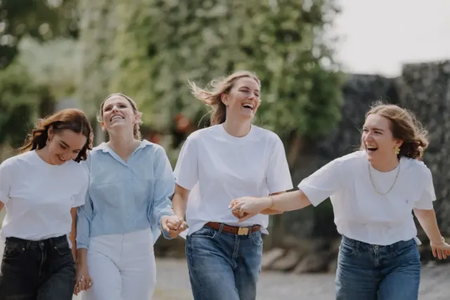 Quatre amies en t-shirt blanc qui rient aux éclats, main dans la main, en plein mouvement
