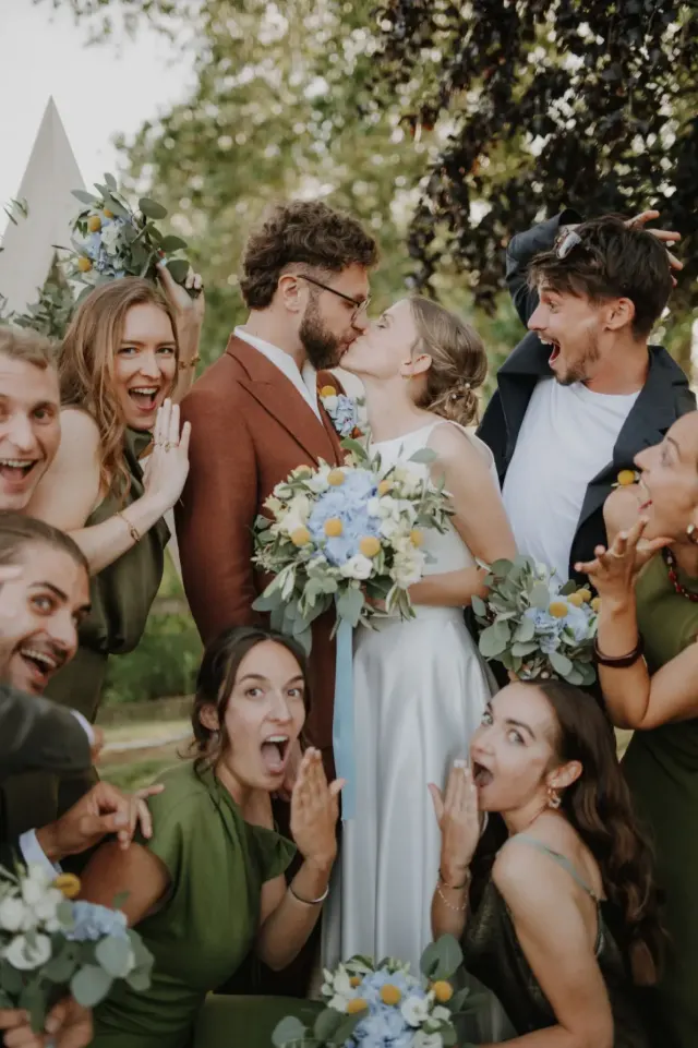Mariés qui s'embrassent entourés de leurs proches hilares et grimaçants, bouquets de fleurs bleues et jaunes